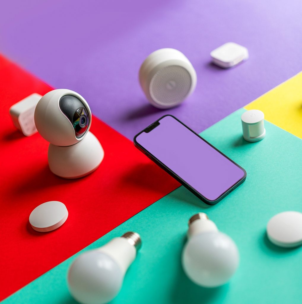 Wireless smart home devices surrounding a smartphone with a blank purple screen on a background of red, purple, teal, and yellow color squares